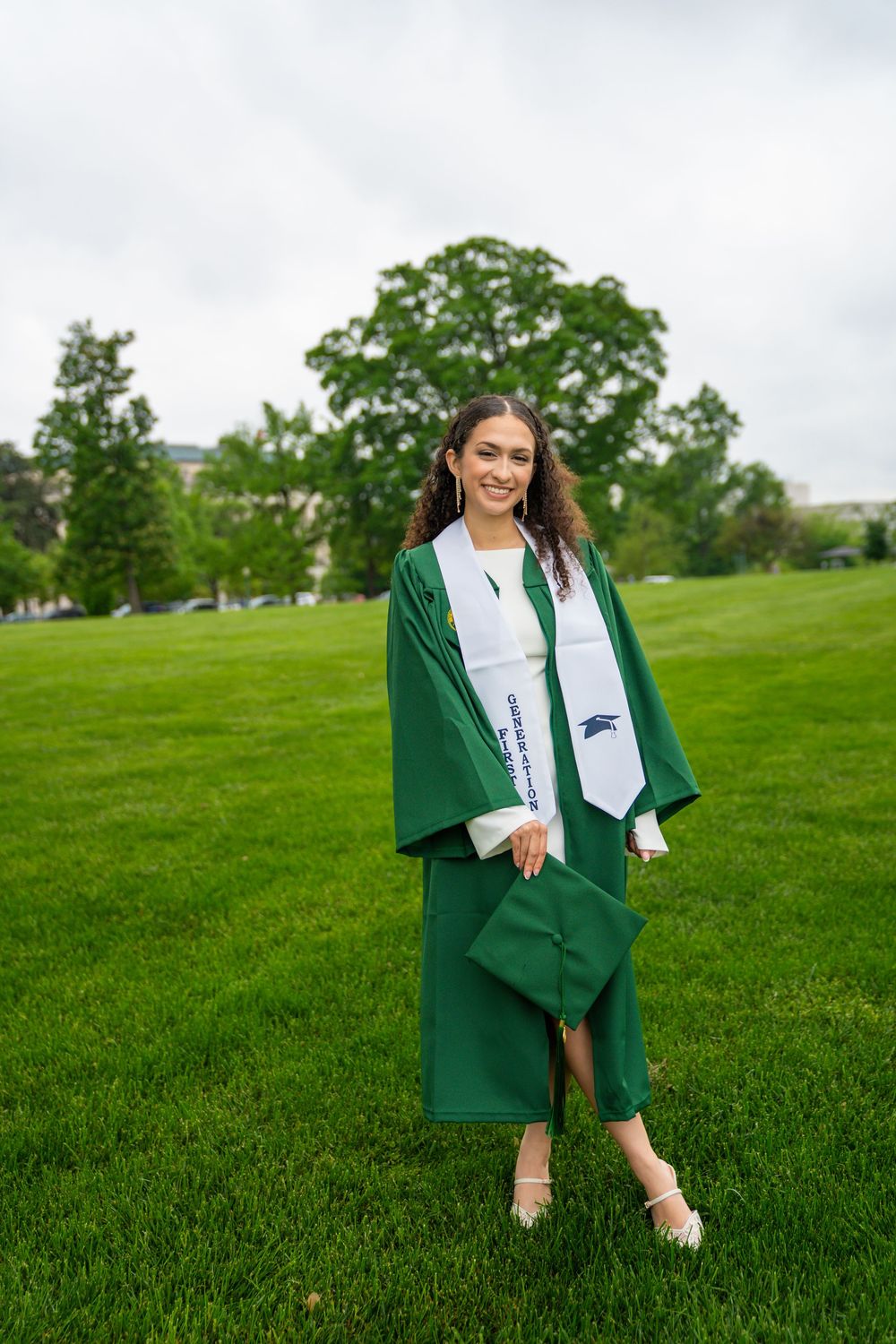 Graduate in green cap and gown poses on grassy lawn with trees in background.