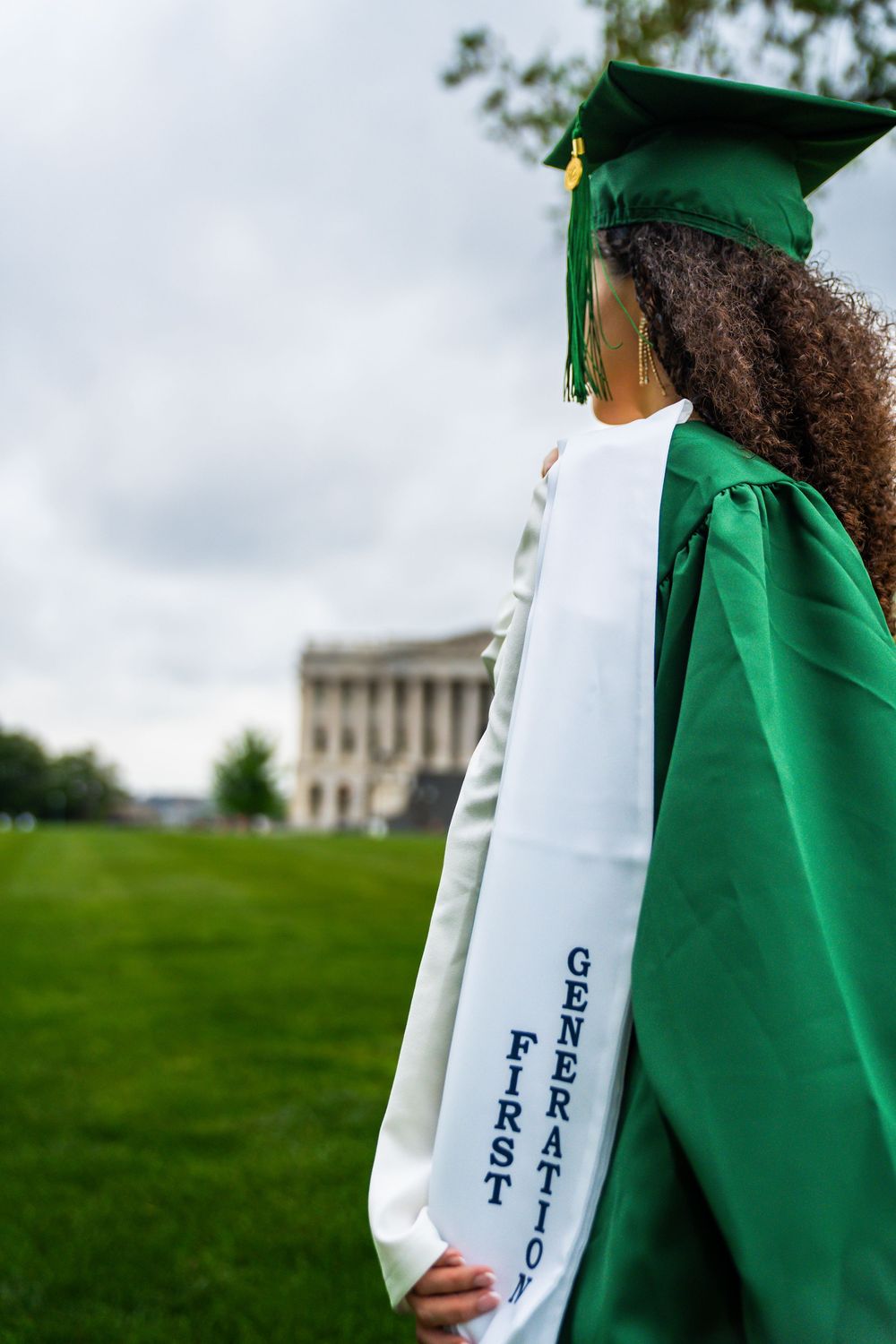 Detail shot of graduation stole and green gown against cloudy sky with building in background.