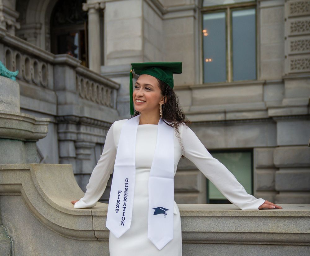 Graduate in white dress and green cap poses on stone steps of academic building.