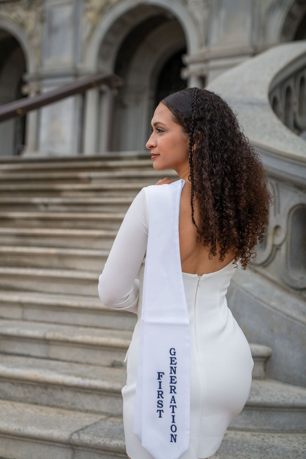 Back view of graduate in white dress and stole standing on ornate stone staircase.