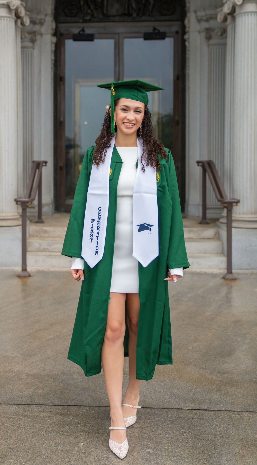 Graduate in green cap and gown with white dress poses on steps of classical building.