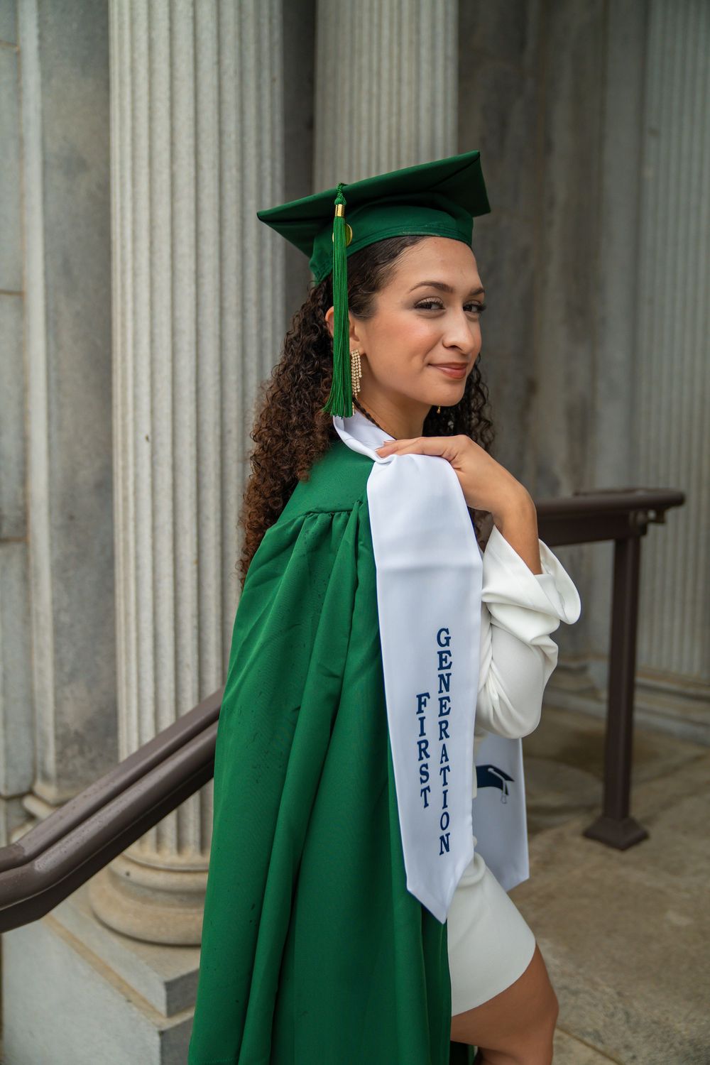 Graduate in white dress and green cap and gown poses elegantly against classical stone columns.