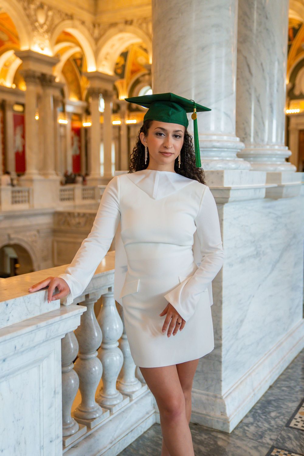 Graduate in white dress and green cap poses in marble hallway with ornate architecture.