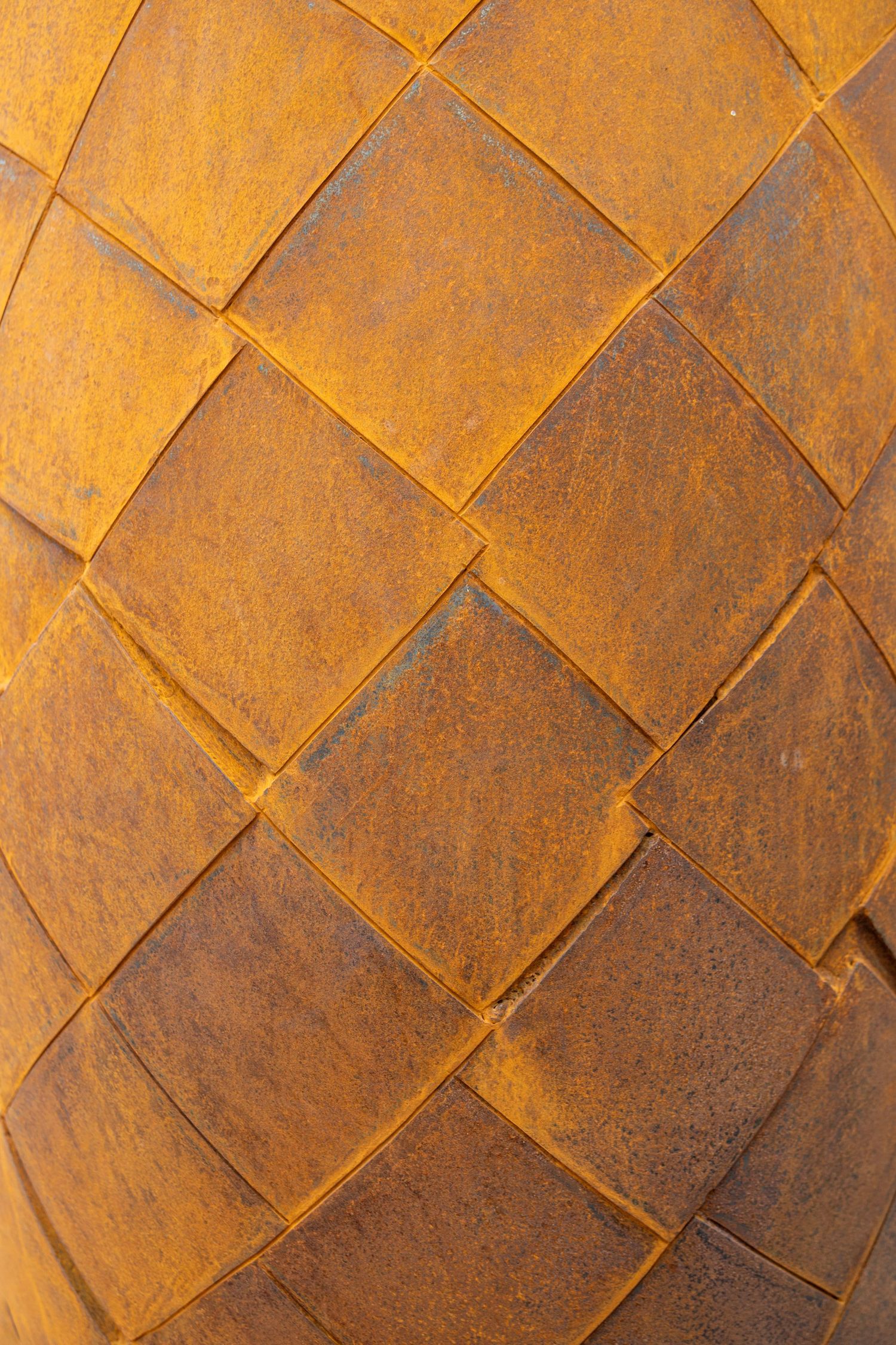Close-up of a weathered Corten steel surface with a diagonal woven diamond pattern in warm rust tones.