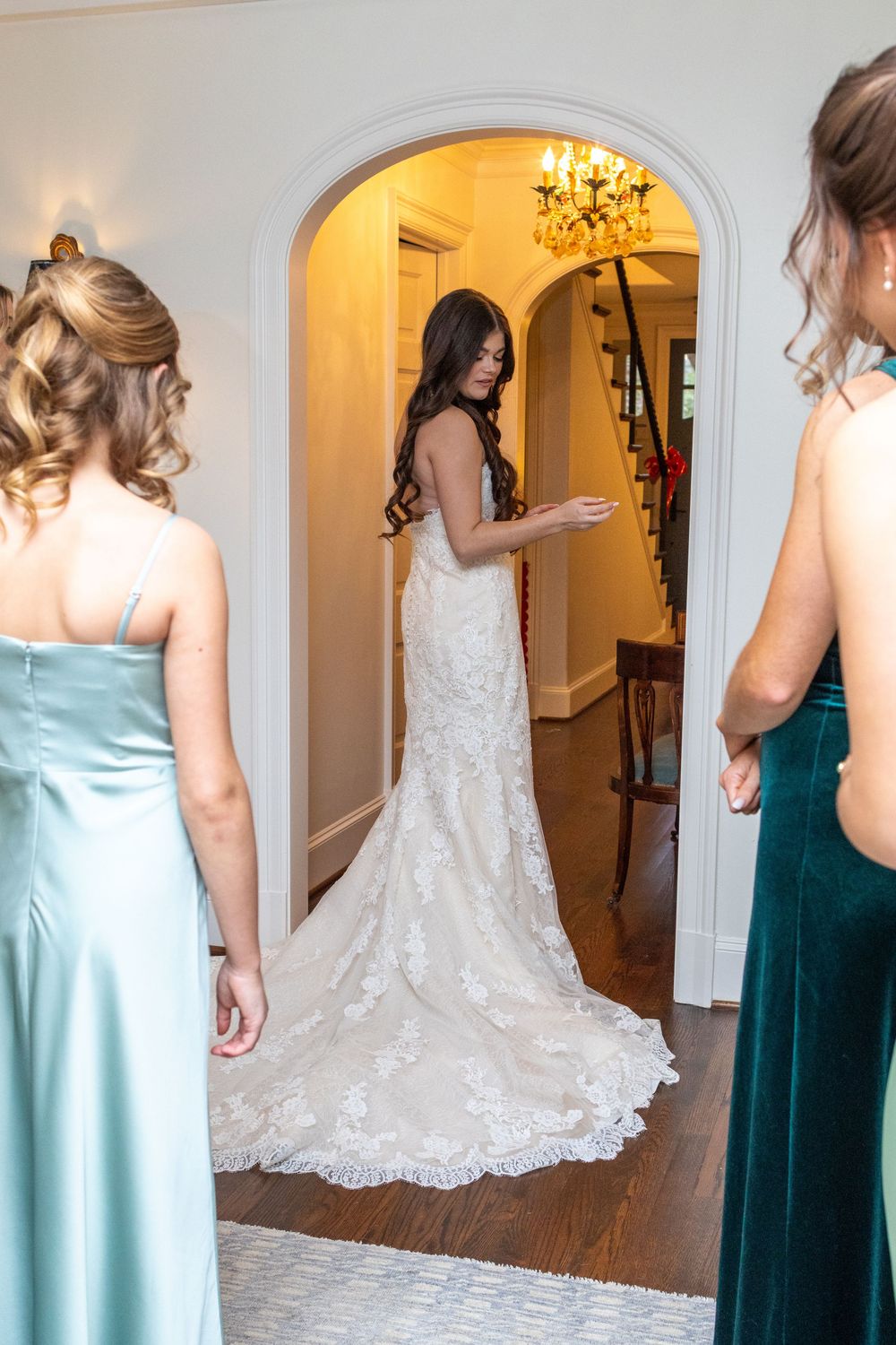 Bridesmaids in mint and emerald dresses help with a white lace wedding gown in a hallway.