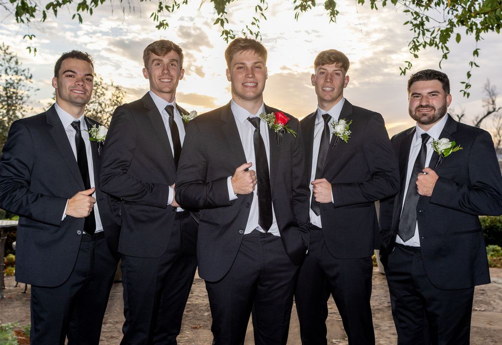A group of five groomsmen in black suits with boutonnieres pose together outdoors at a wedding venue.