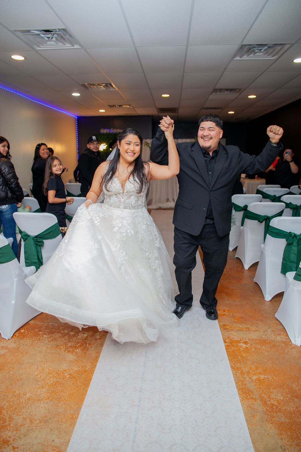 Wedding party members dancing and celebrating in a reception hall with white chairs.