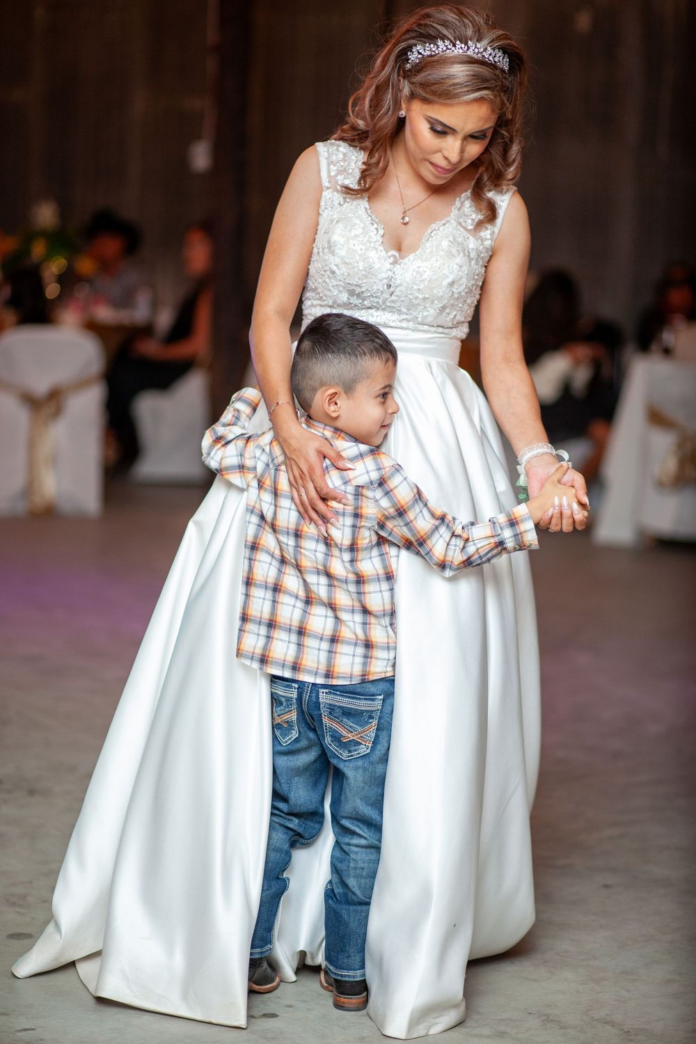 A young child in plaid and jeans dances with someone in a white wedding dress at a reception.