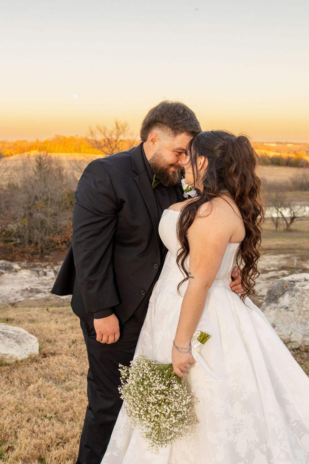 A romantic couple shares tender moments at sunset in a rustic outdoor setting with baby's breath bouquet.