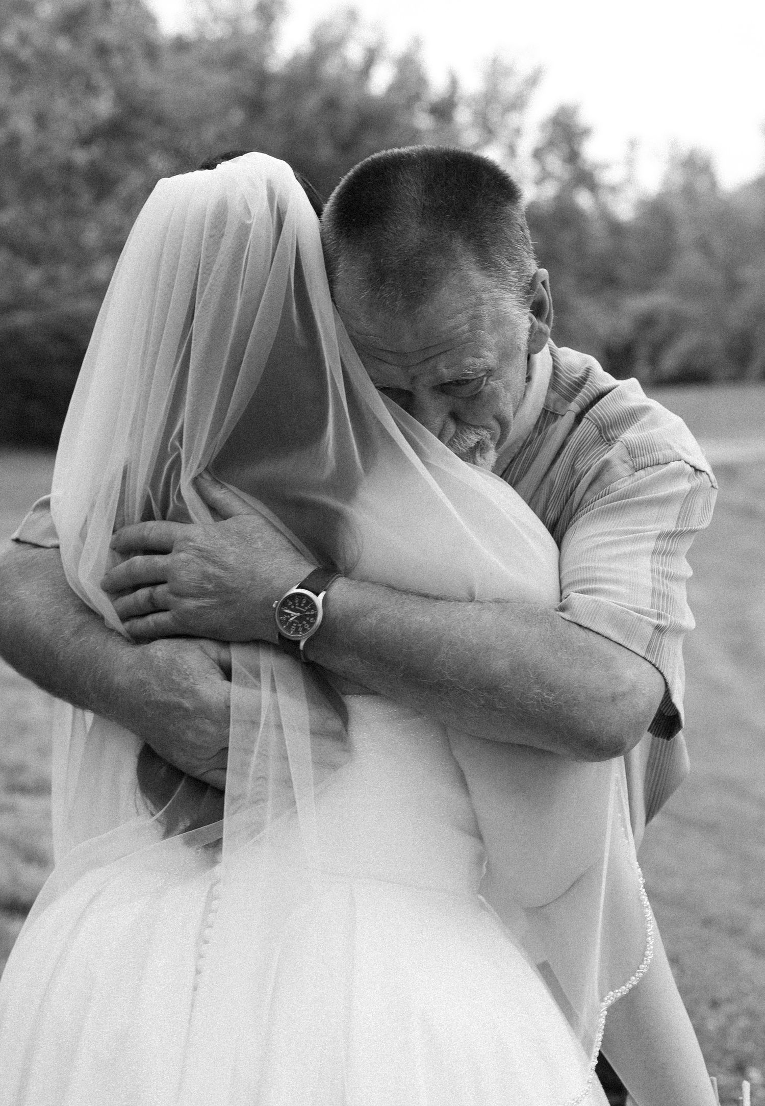 A tender black and white wedding moment captured outdoors as a father embraces his daughter in her bridal gown and veil.