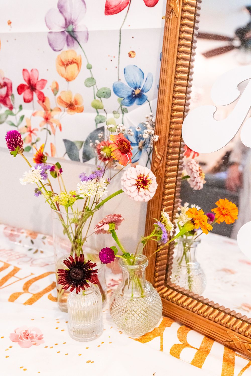Clear glass vases with colorful wildflowers sit beside a gold-framed mirror with floral wallpaper background. Baby Shower decor close-up