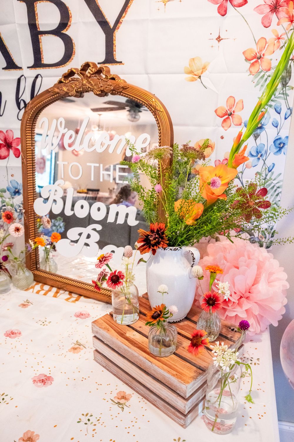 A vintage mirror with 'Welcome to the Bloom' text and floral decor arrangement featuring gladiolus and pink peonies. Baby Shower decor