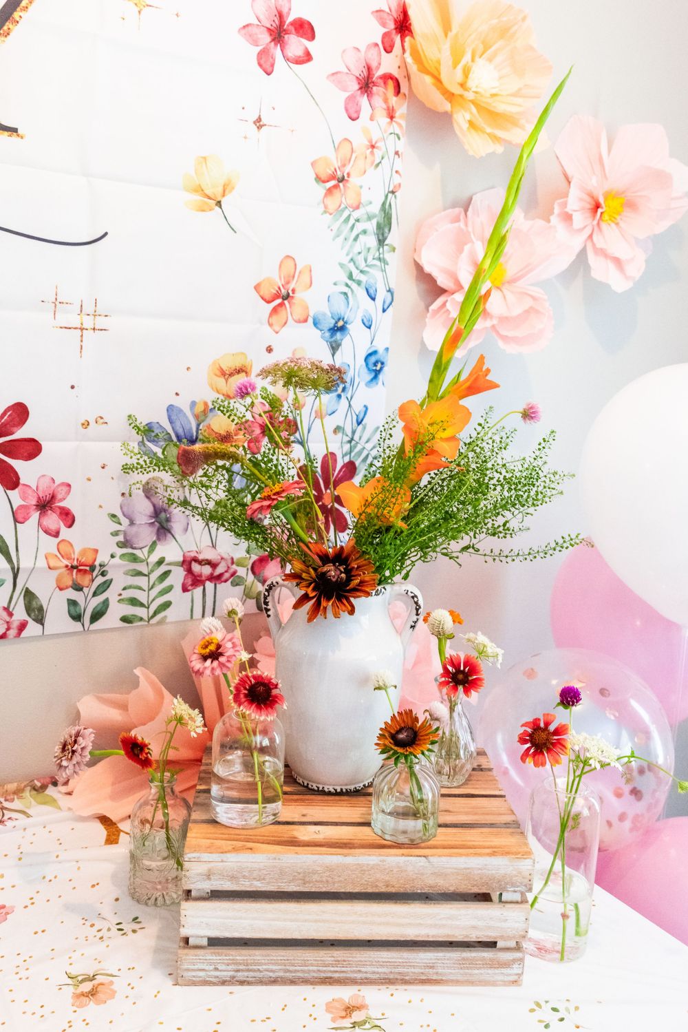 Colorful floral arrangement with pink and yellow blooms in white vase against floral wallpaper. Baby Shower Decor