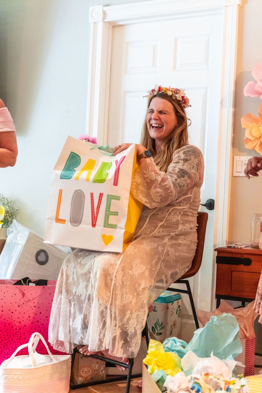 A joyful baby shower celebration with a person in a lace dress laughing while holding a "Baby Love" gift bag.