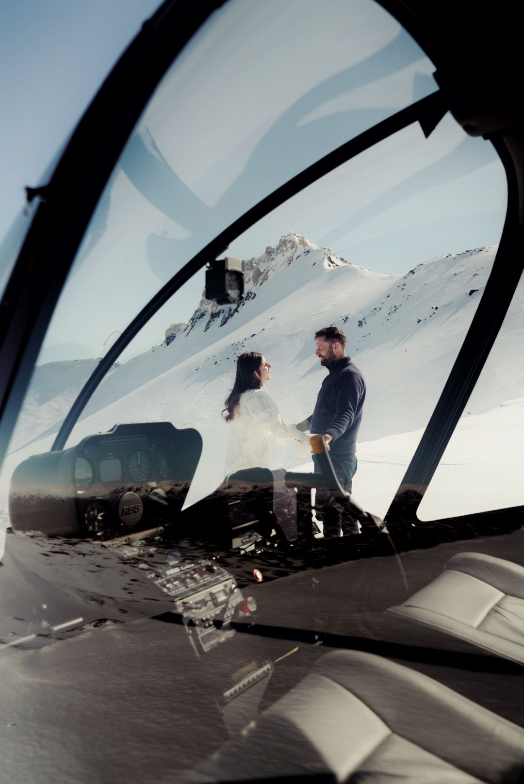 View from inside a helicopter cockpit overlooking snowy mountain peaks.