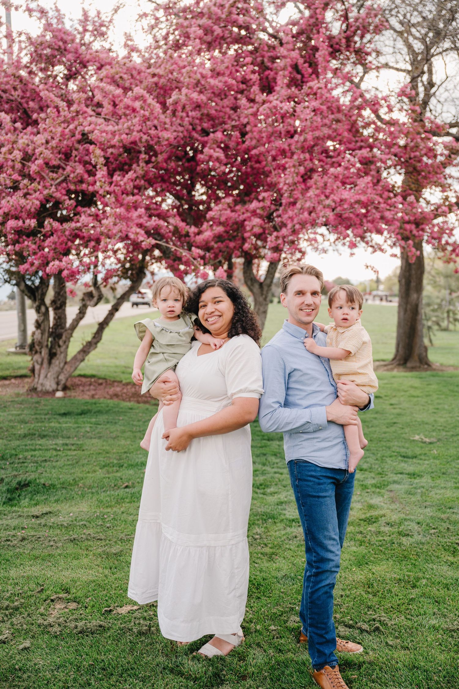 A family poses together under a blooming pink cherry blossom tree in a spring park setting.