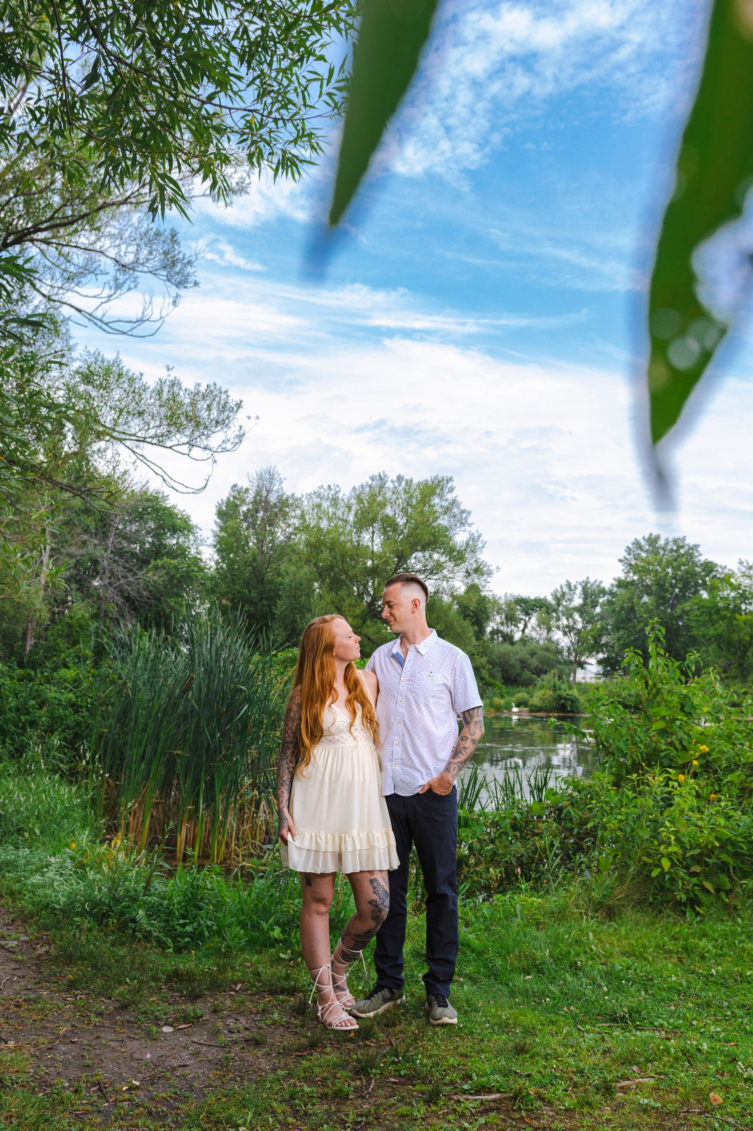 Romantic couple standing by a pond surrounded by lush greenery on a sunny summer day.