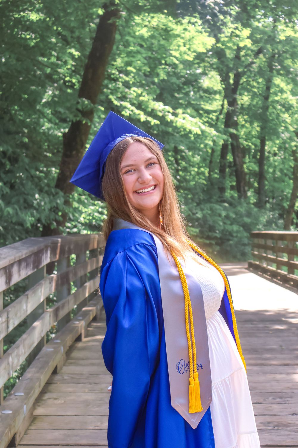 Graduate in blue cap and gown poses on wooden boardwalk surrounded by green trees.