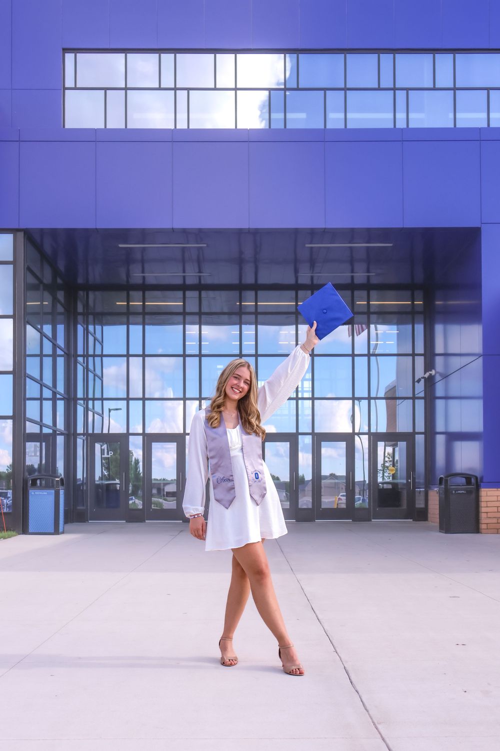 Graduate in white dress celebrating with blue cap raised outside modern glass building.