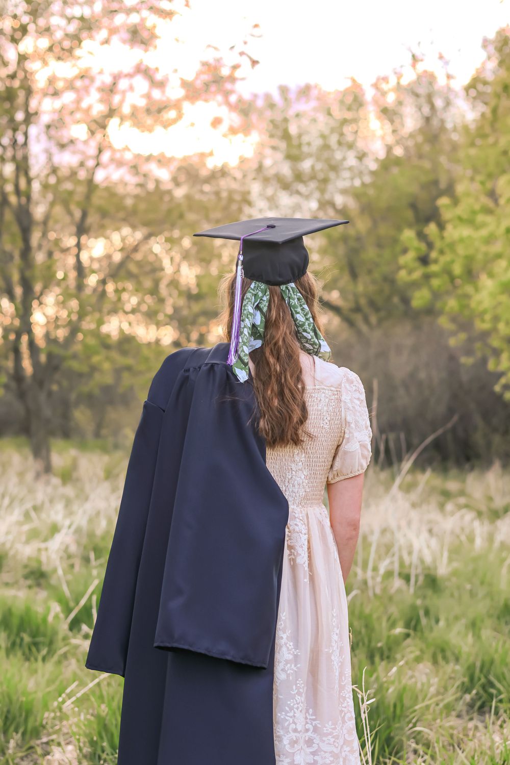 Back view of graduate wearing cap and gown with lace dress in natural outdoor setting.