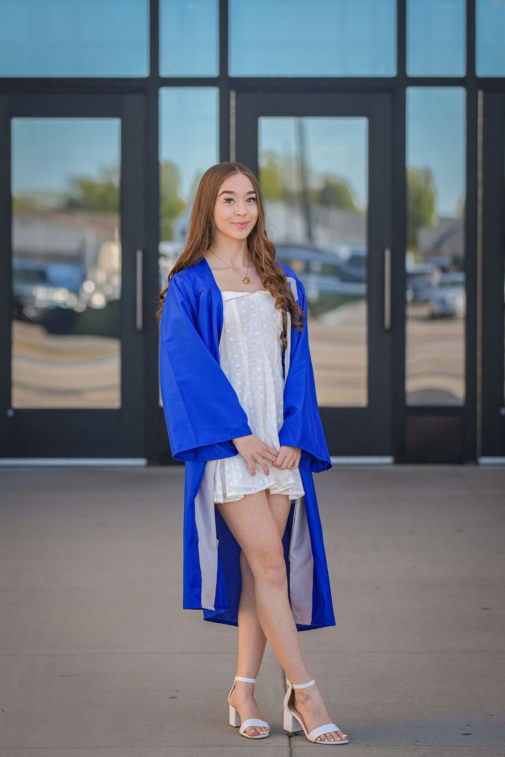 Young woman in blue graduation gown over white dress poses confidently outside school entrance in heels.