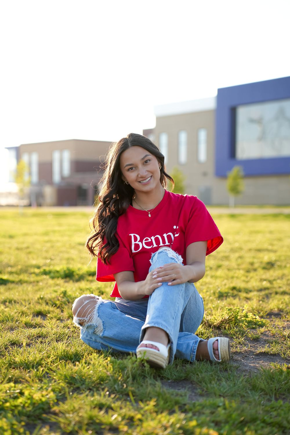 Student wearing a red Benny t-shirt and ripped jeans sits on grass during golden hour near campus buildings.