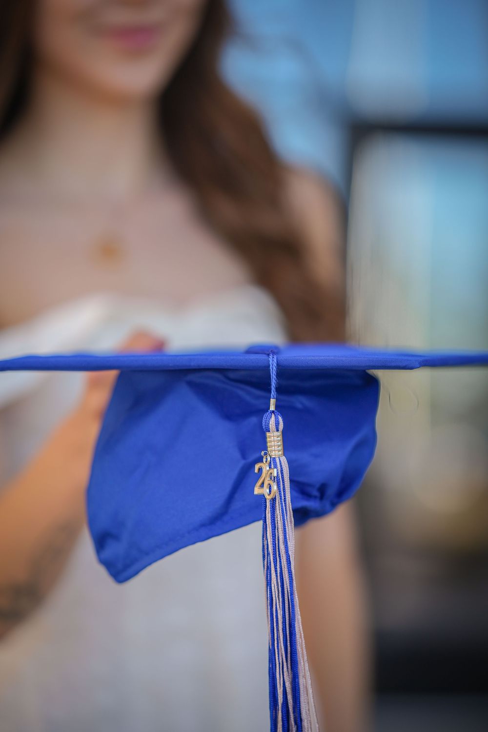 A blue graduation cap with tassel held in front of a woman in a white dress.
