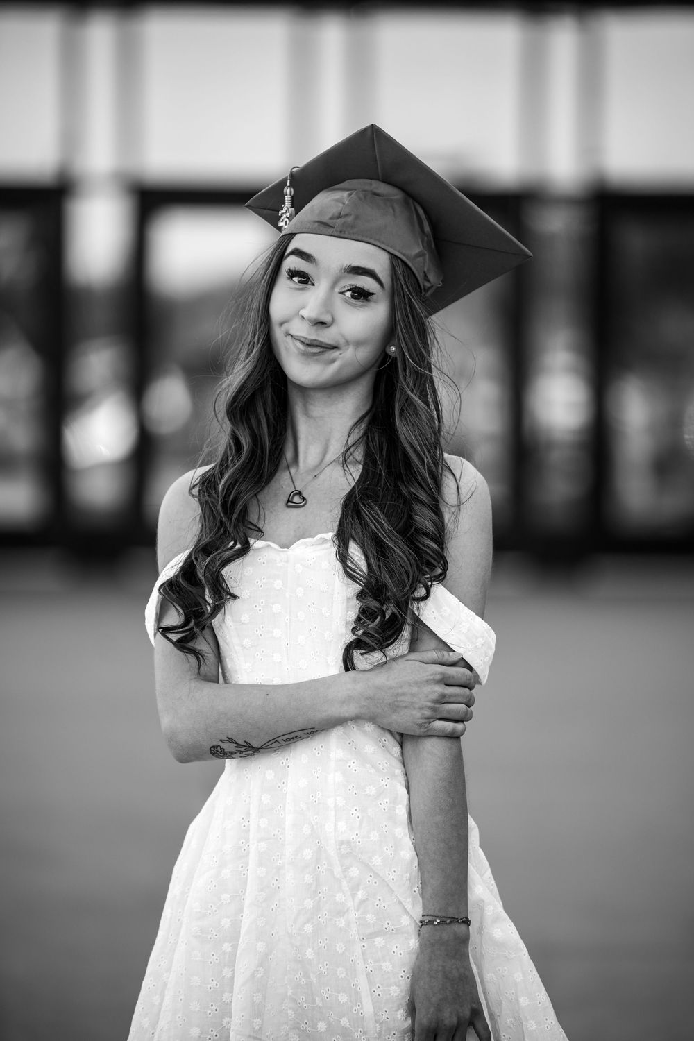 Black and white portrait of a young woman in graduation cap and white dress holding diploma outdoors.