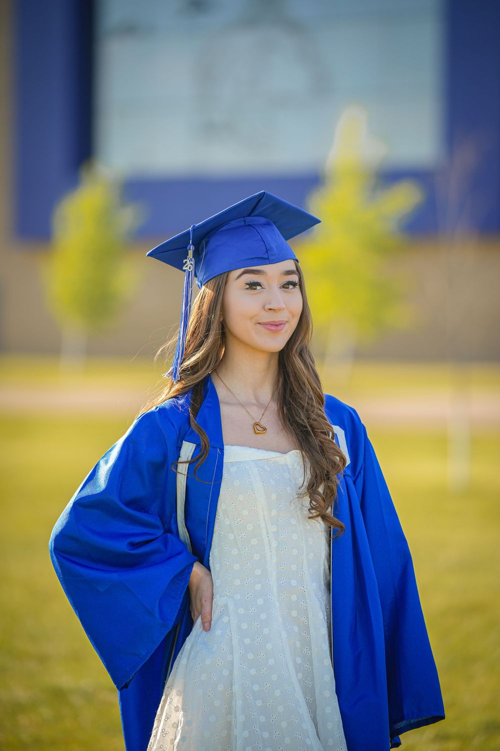 Young woman in blue graduation cap and gown smiling outdoors on a sunny day.