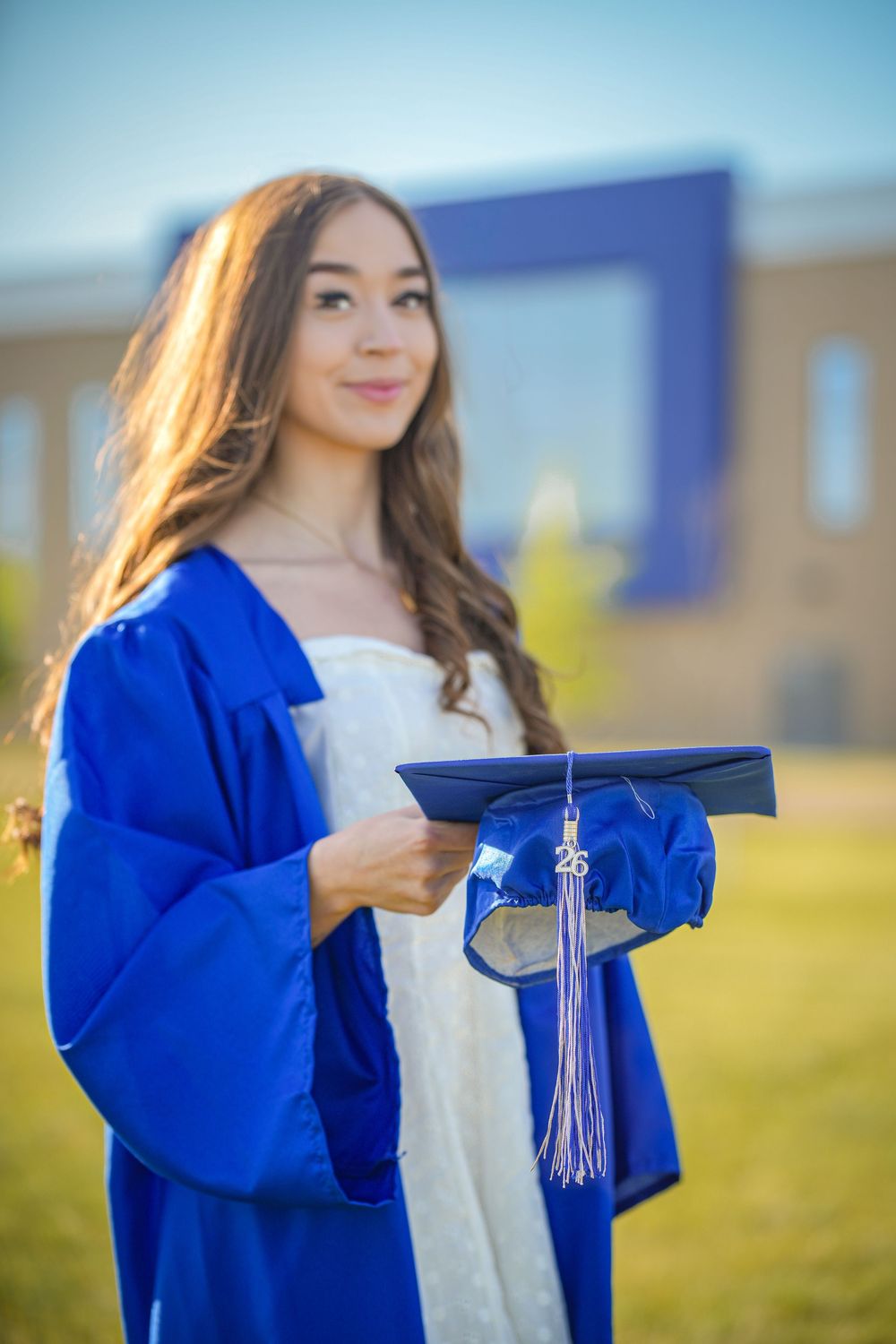 Young woman in blue graduation cap and gown holding her mortarboard outdoors near school building on a sunny day.