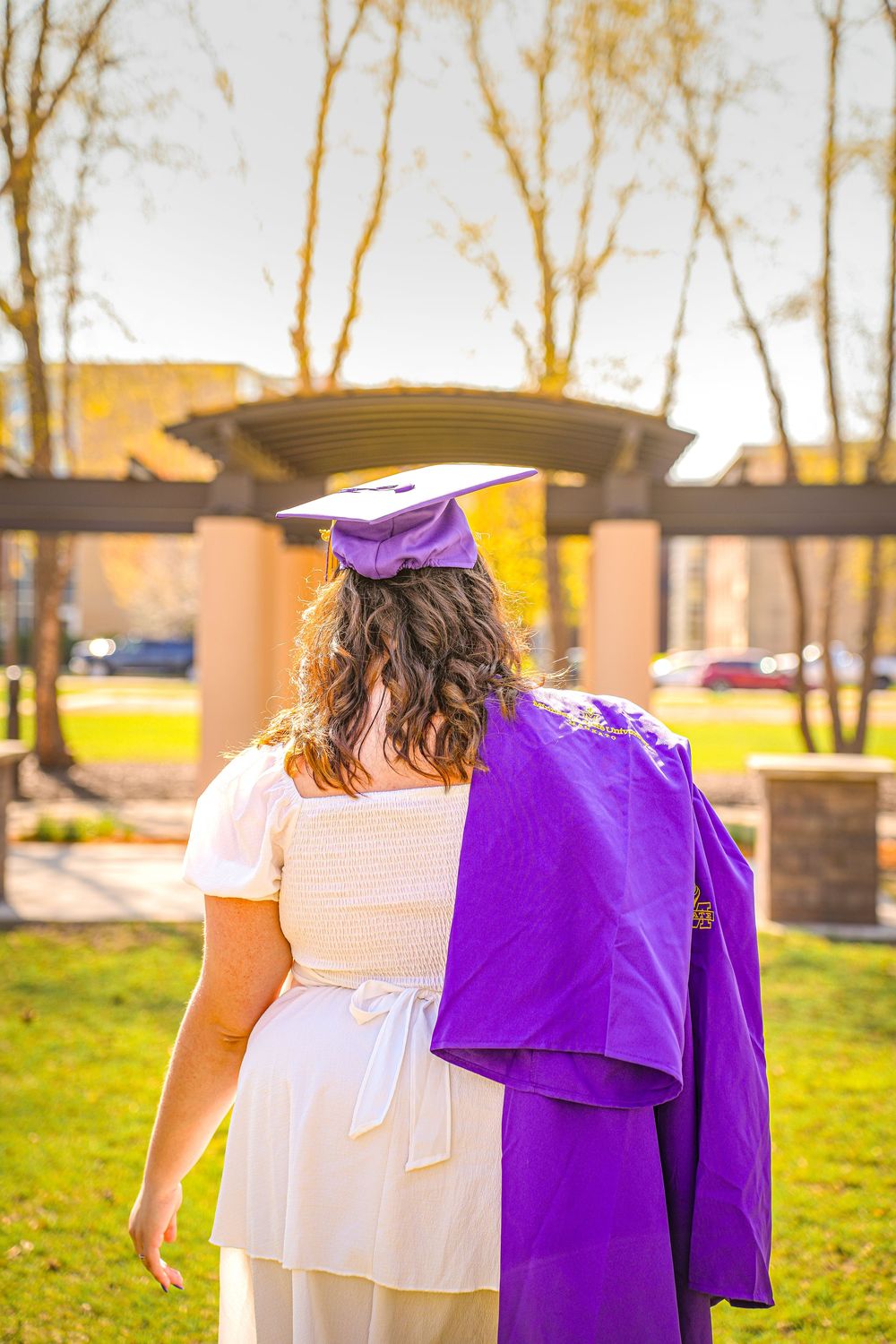 A graduate stands holding their purple graduation gown and wearing a matching cap in a white dress outdoors at sunset.
