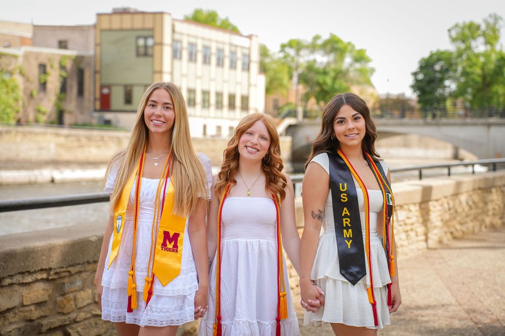 Three graduates in white dresses and graduation stoles stand together smiling outdoors on a sunny day.