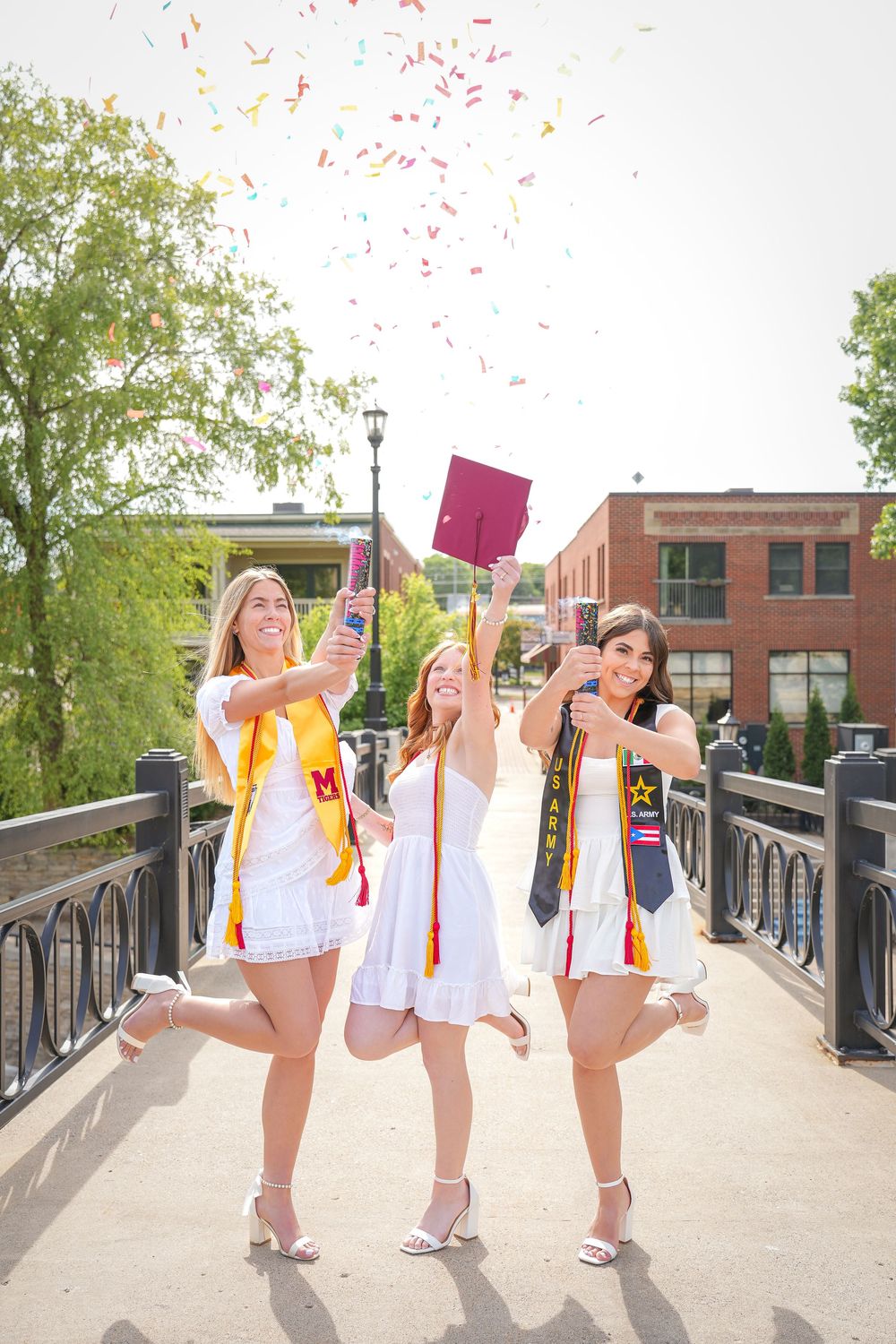 Three graduates in white dresses and honor cords celebrate with confetti and raised diplomas on a bridge walkway.