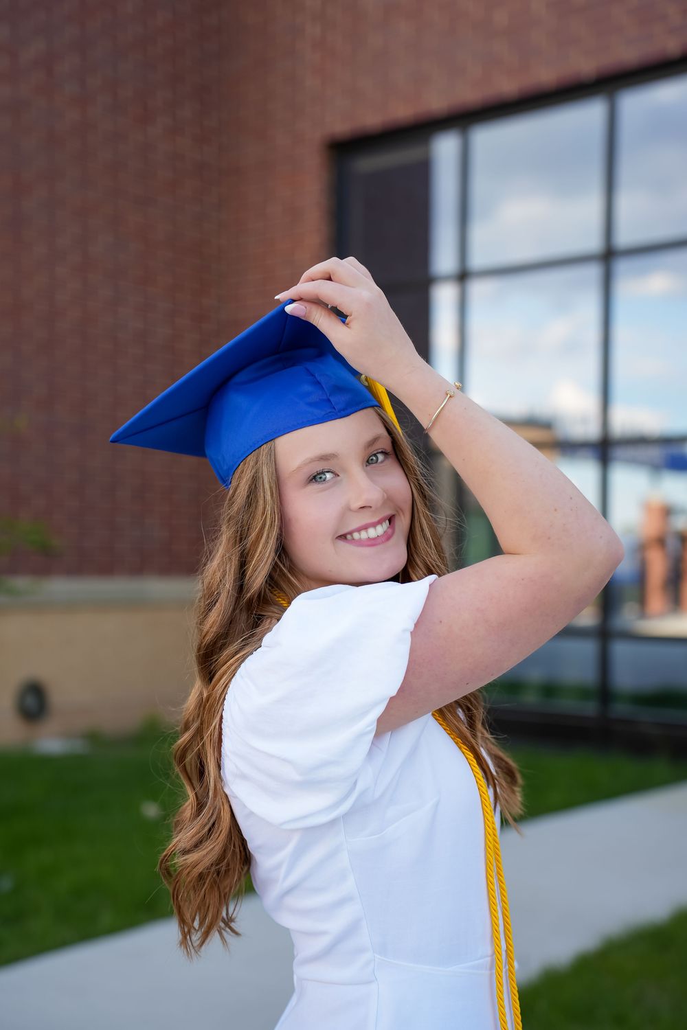 Graduate in blue cap and white dress poses playfully outdoors near a brick building.
