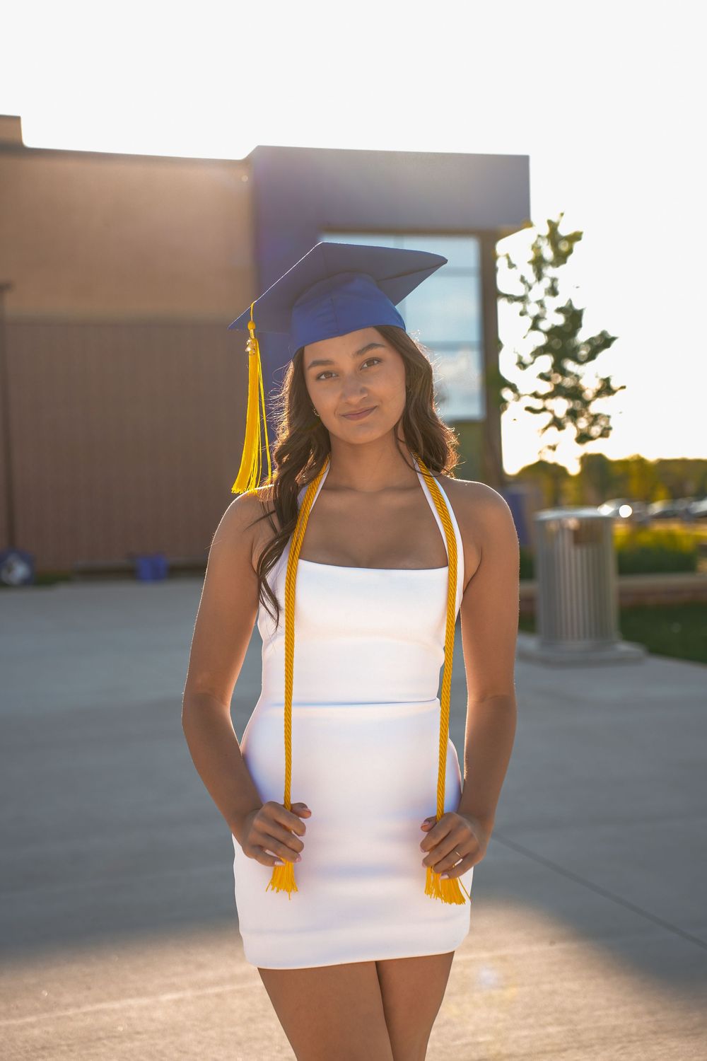 Graduate in white dress and blue cap poses in golden sunset light outside school building.