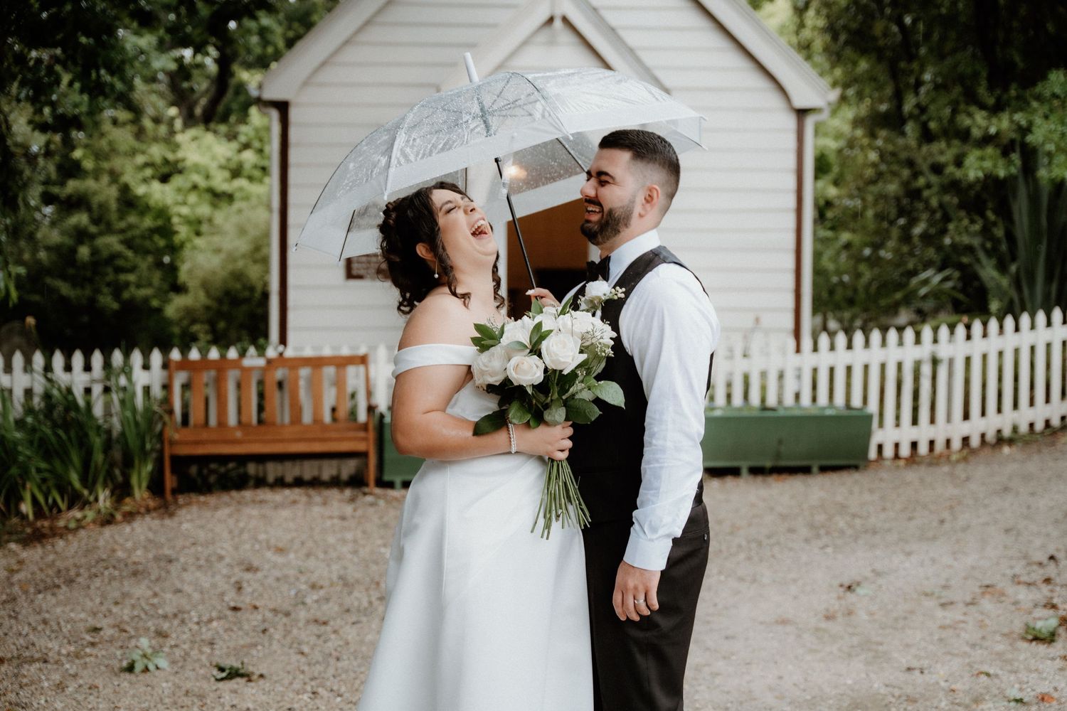 Wedding couple shares joyful moment under umbrella near white chapel with picket fence.