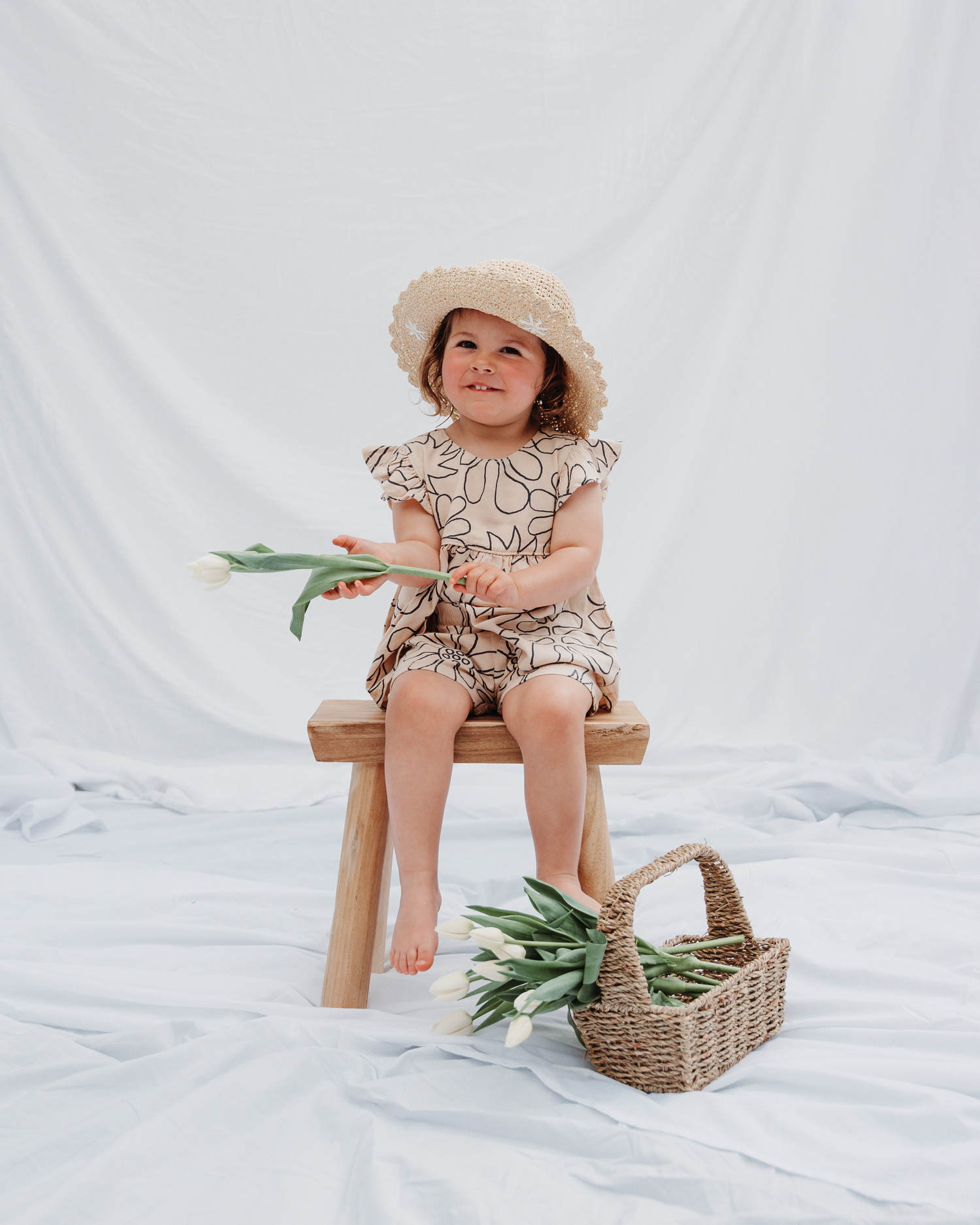 A child in a patterned outfit sits on a wooden bench with flowers and a woven basket.