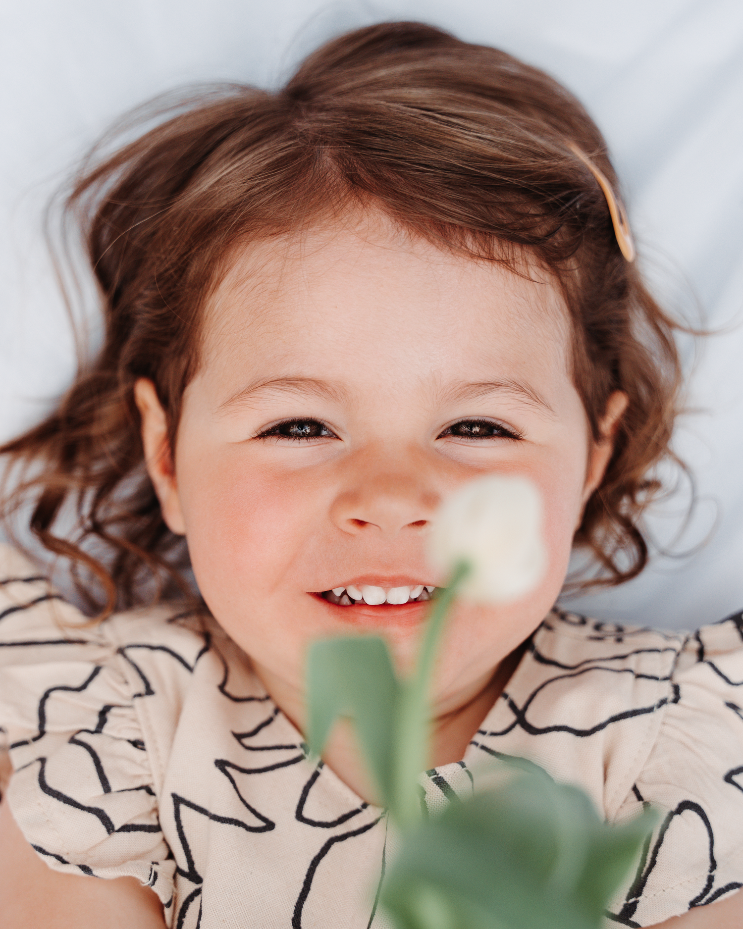 A close-up portrait of a smiling child in a patterned dress with a plant in soft focus.