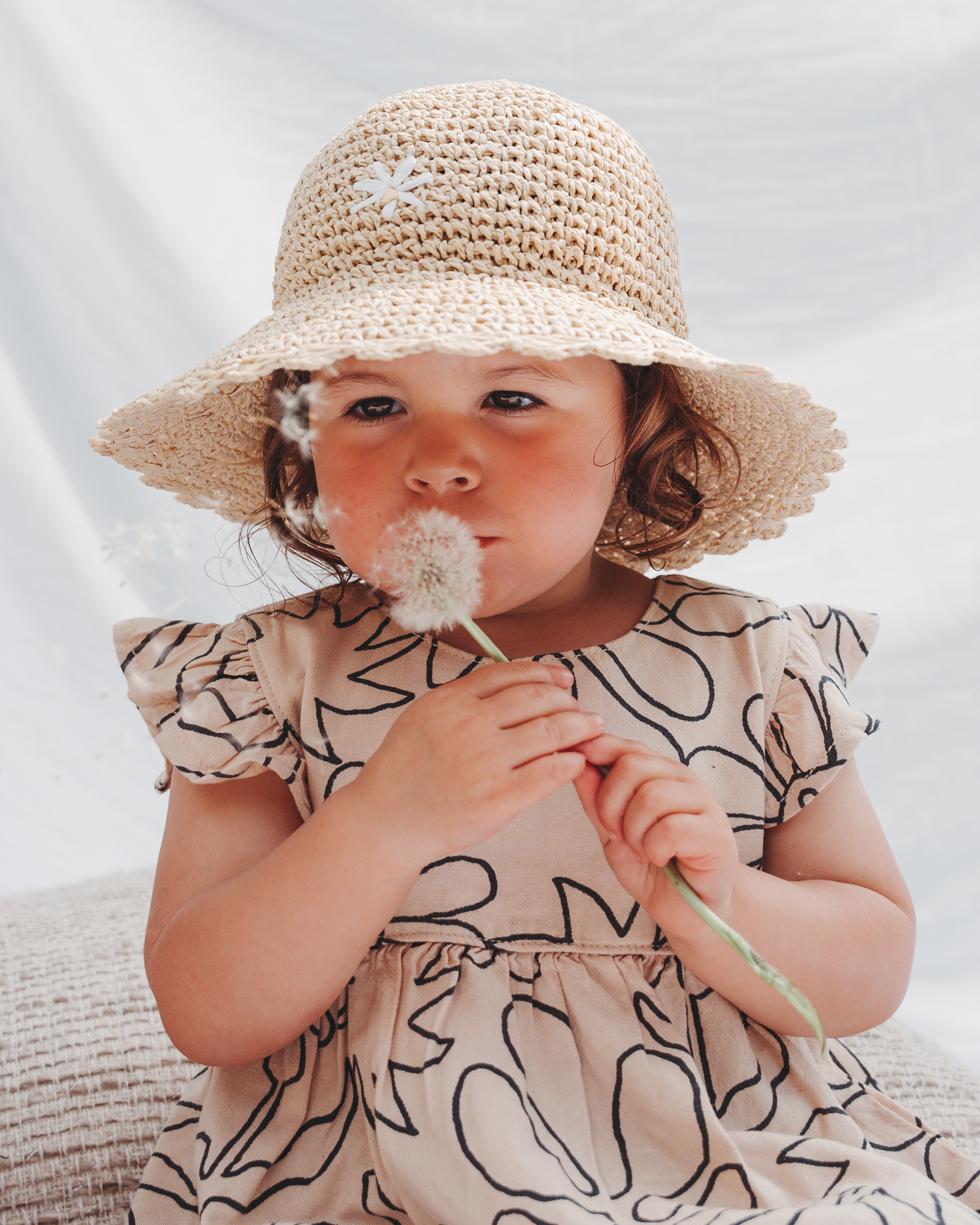 A child in a patterned dress and straw hat blows on a dandelion against a white backdrop.