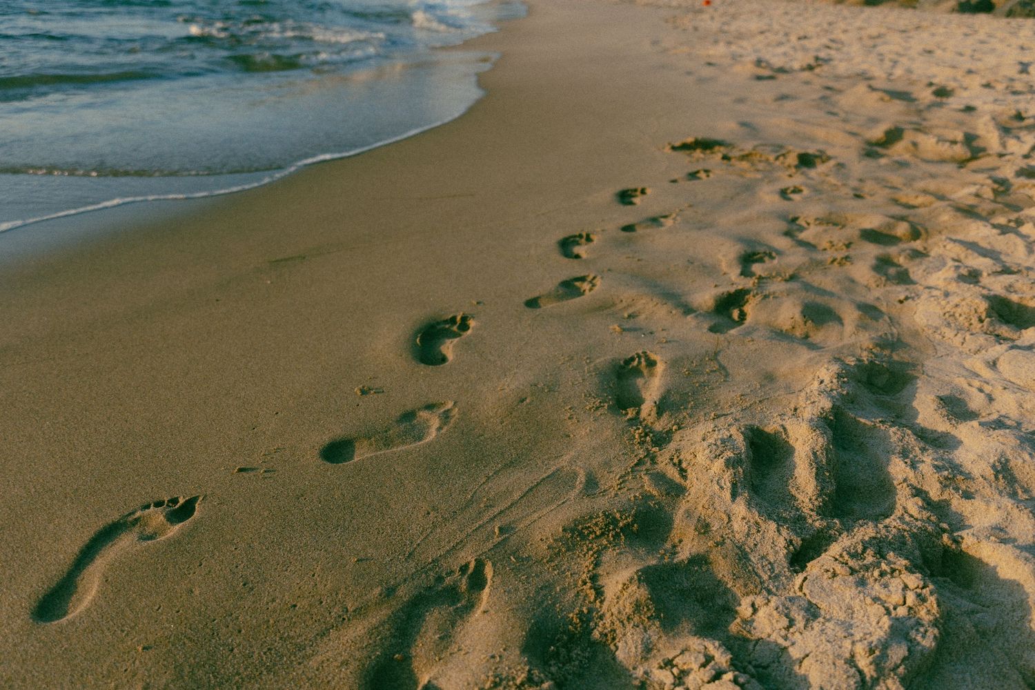 Multiple footprints in wet sand along a beach shoreline with gentle waves rolling in at sunset.