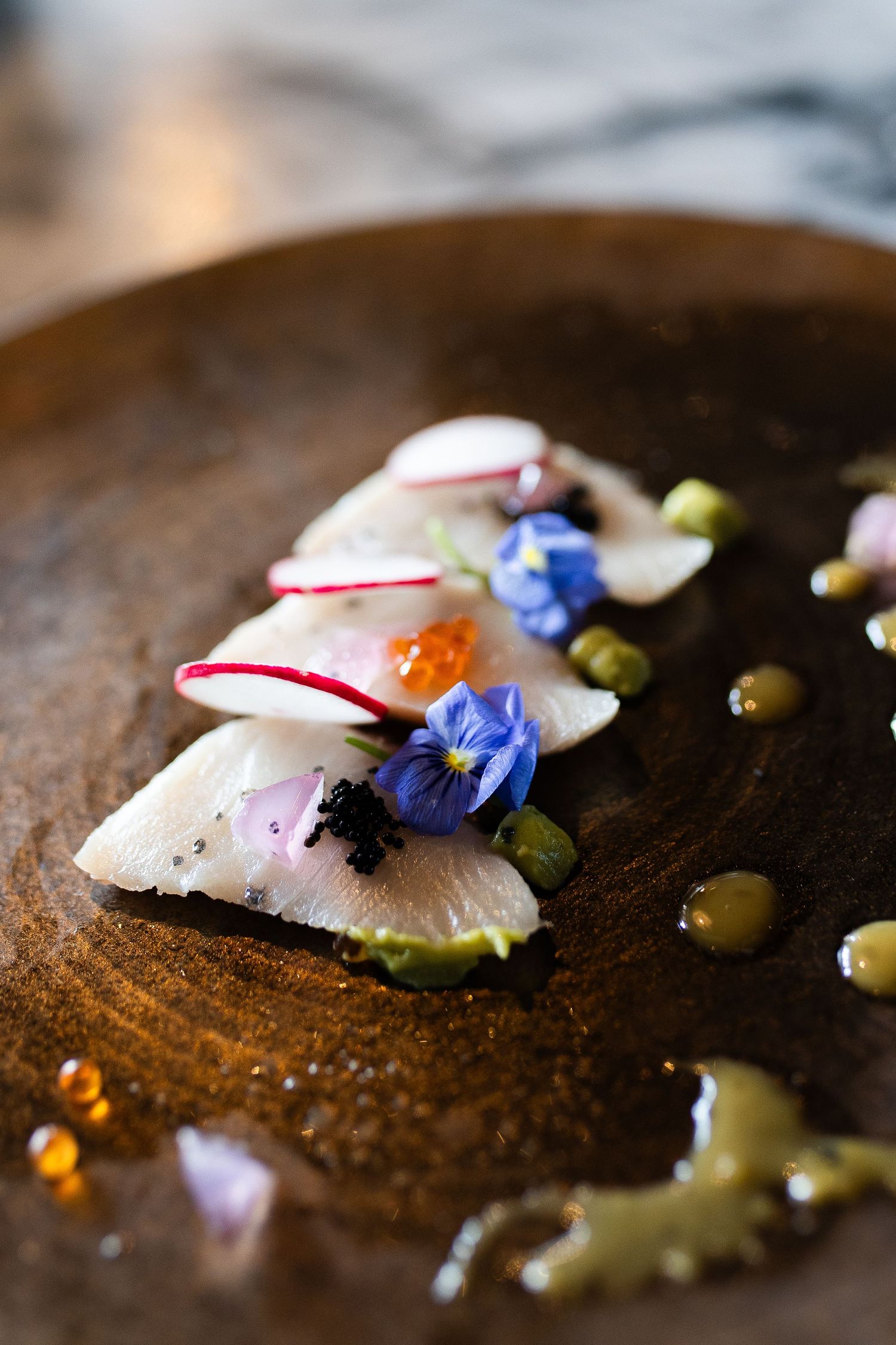 Elegant raw fish dish garnished with thinly sliced radishes, vibrant blue edible flowers, and dots of sauce on rustic plate.