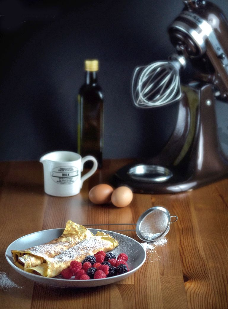 A freshly made crepe with powdered sugar and berries next to eggs and a stand mixer on a wooden table.