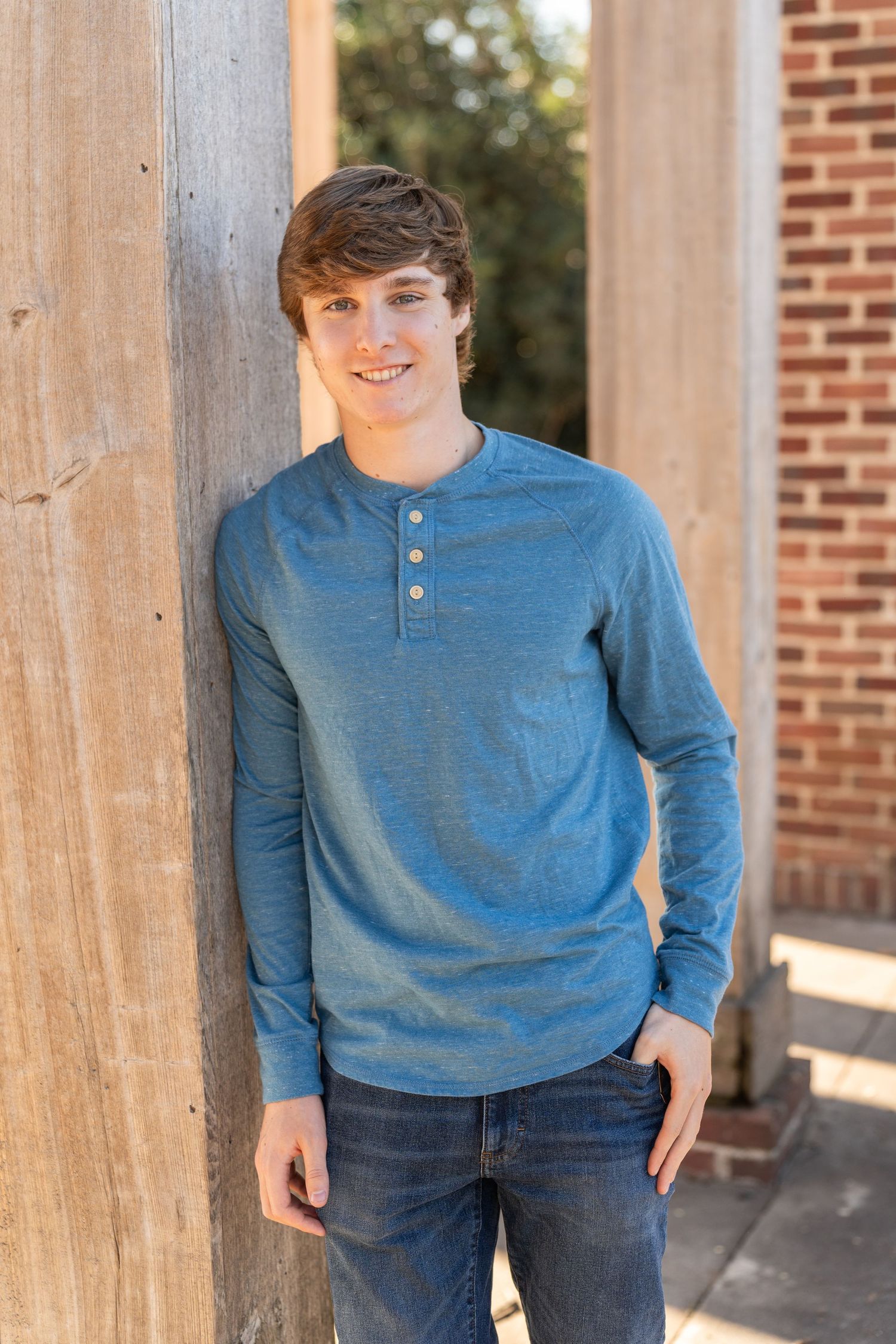 Young person in blue long sleeve henley shirt and jeans leaning against wooden post outdoors by brick building.