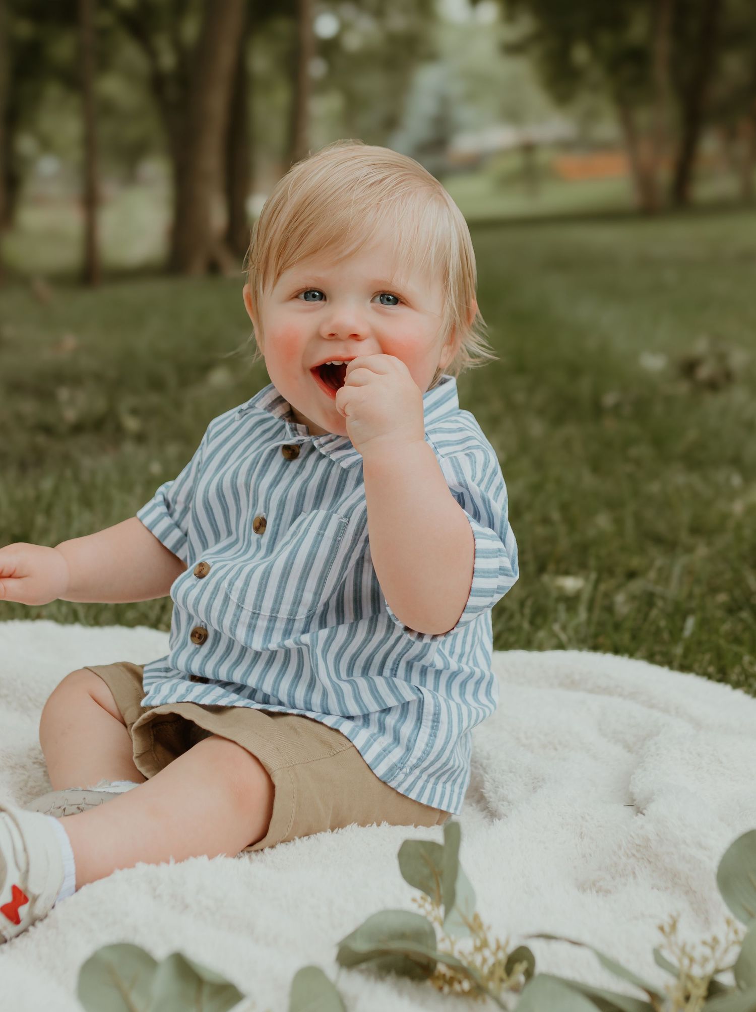 Small child in striped shirt and shorts sits on white blanket outdoors while smiling and playing in a grassy area.
