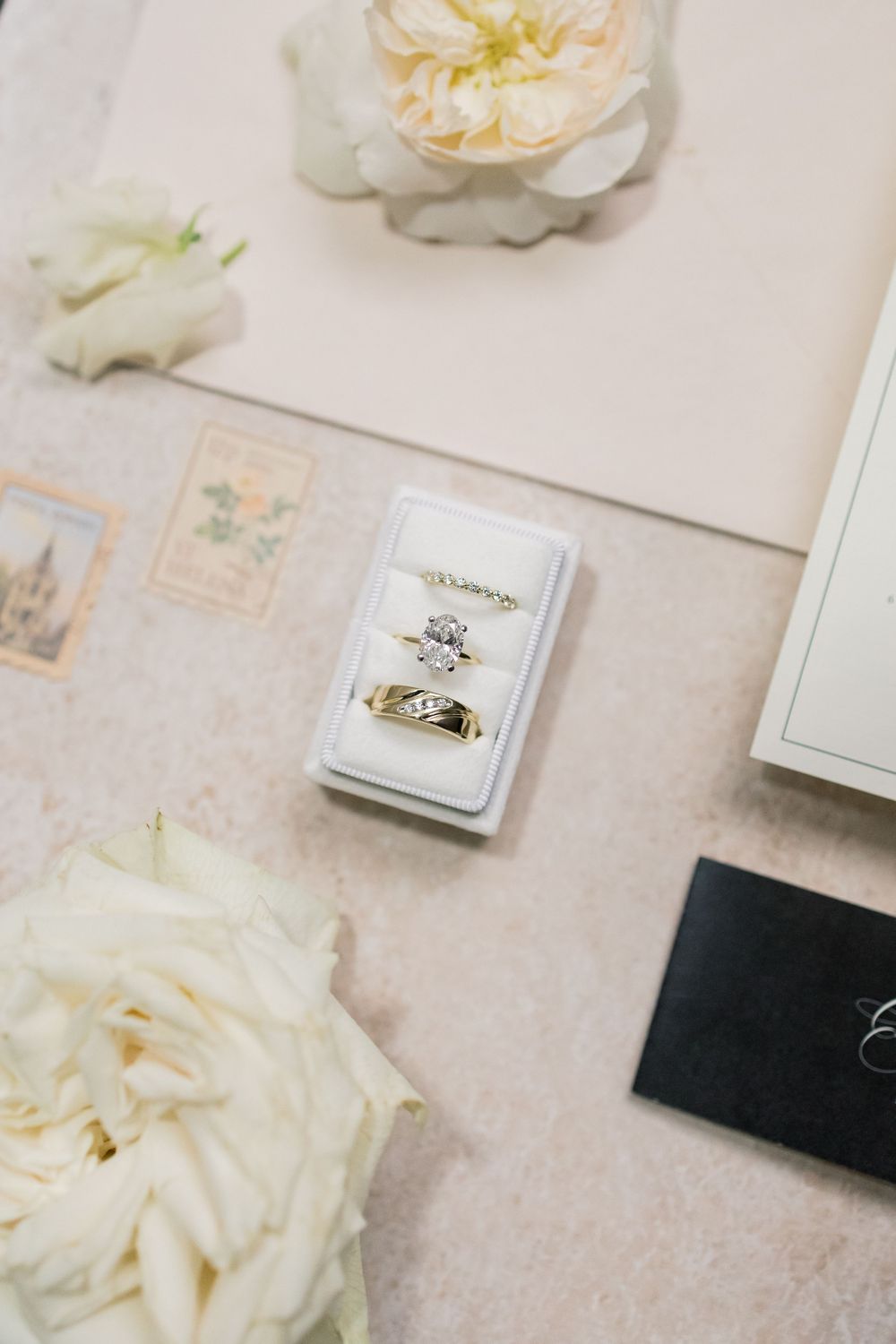 Wedding ring displayed in a white jewelry box surrounded by delicate cream roses and vintage stamps on a light surface.