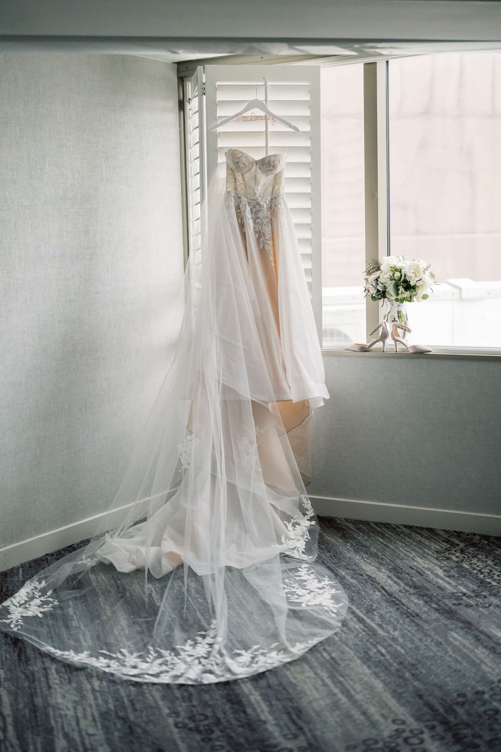 An elegant white wedding dress and veil hanging in a window with soft natural light and floral decor.