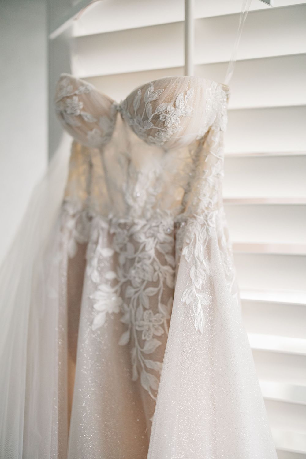 Elegant white wedding dress with delicate lace details and strapless bodice hanging by window blinds for bridal preparation.
