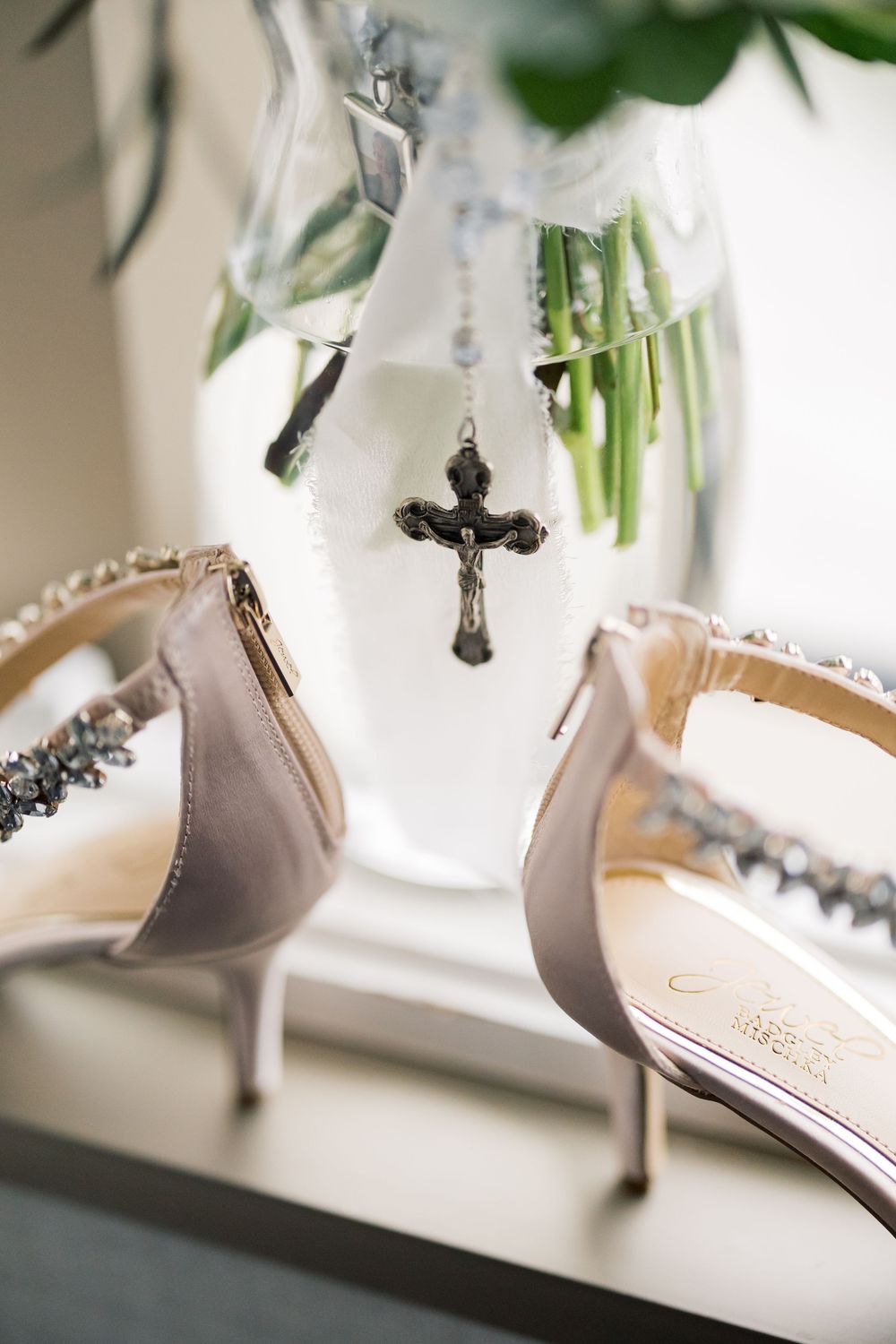 Elegant gray heels with crystal details sit beside a delicate cross pendant and glass vase with greenery.