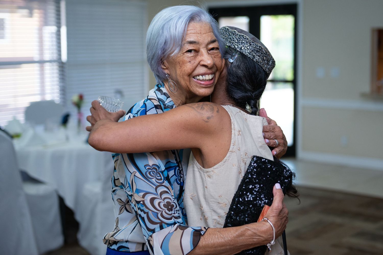 Two women share a warm embrace at an indoor event venue decorated for a celebration.
