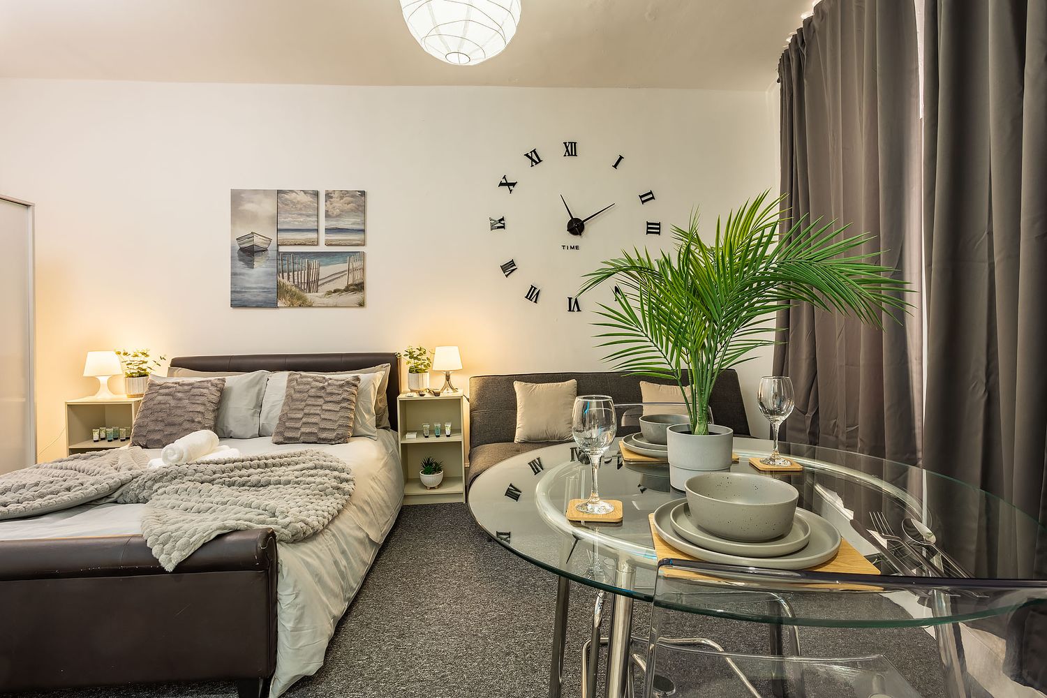 Modern hotel room interior with grey bedding, glass dining table, potted palm plant and decorative wall clock.